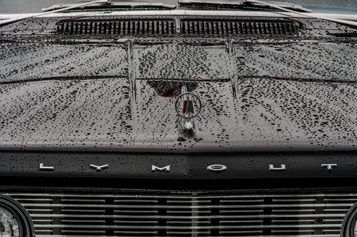 Black and white close view of a 1960s Plymouth head on in the rain, the hood covered in standing water beads, the chrome hood ornament centered above the PLYMOUTH letters and the full width of the bar grille below.