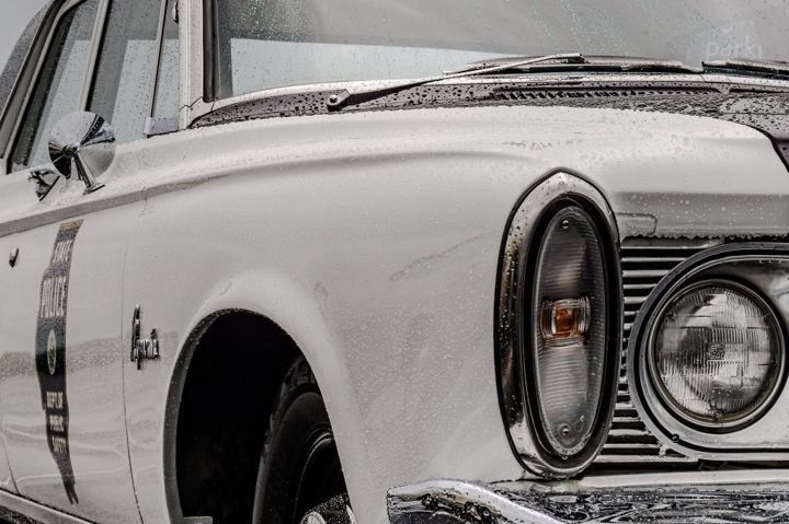 The front fender of a restored white Illinois State Police Plymouth beaded with rain, its stacked headlight and turn lamp at right and the State Police door lettering running down the left of the frame under a spotlight mirror.