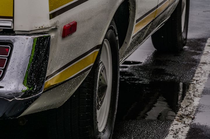 Low three quarter view of a white muscle car with gold and black side stripes parked on soaked pavement, its rear wheel and dented quarter panel filling the left of the frame and the car mirrored in the water pooled along a painted parking line.