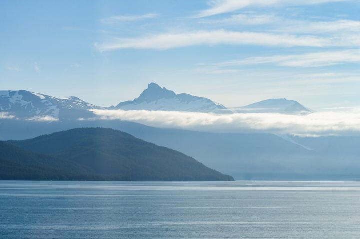 A snow covered peak standing above a flat bank of cloud with a dark forested ridge below it, still blue water running across the bottom of the frame.