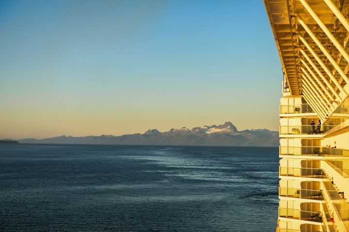 The stacked balconies of a cruise ship curving away in gold light down the right of the frame, with a jagged snow capped coast range across the water on the left.