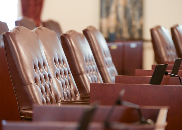 A row of tufted oxblood leather high back chairs behind wooden desks fitted with microphones, in a hearing room with a tapestry on the far wall.