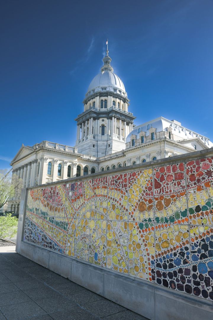 The Illinois State Capitol dome against a deep blue sky, with a long mosaic wall of hand painted tiles in red, yellow, green and blue running along the sidewalk in the foreground.