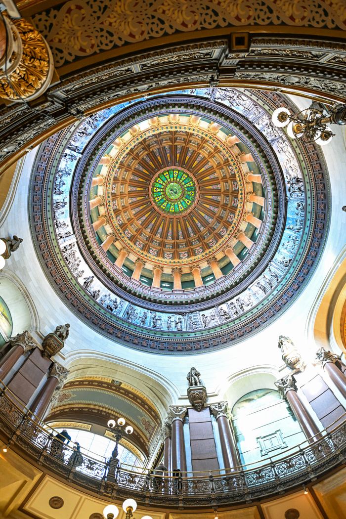 The inside of the Illinois State Capitol dome seen from the rotunda floor, ringed by a painted frieze of figures and rising to a stained glass oculus with the state seal at its center in green and gold.