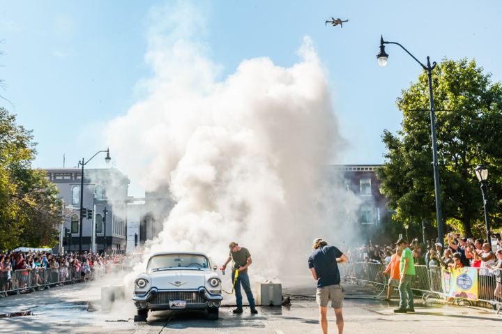 A cream classic Cadillac lit up in a burnout at the center of a closed street, tire smoke towering over the roofline while a crowd watches from behind barricades and a drone hovers overhead.