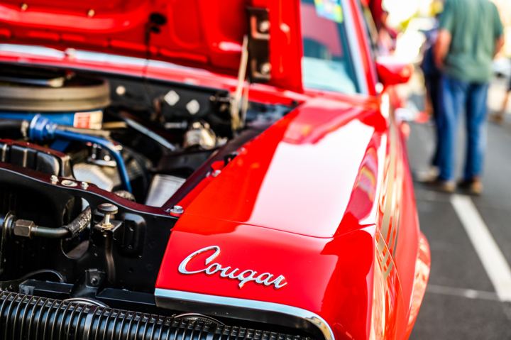 The open hood of a red Mercury Cougar at a car show, blue plug wires and polished chrome under the lid, the Cougar script badge sharp on the front fender.