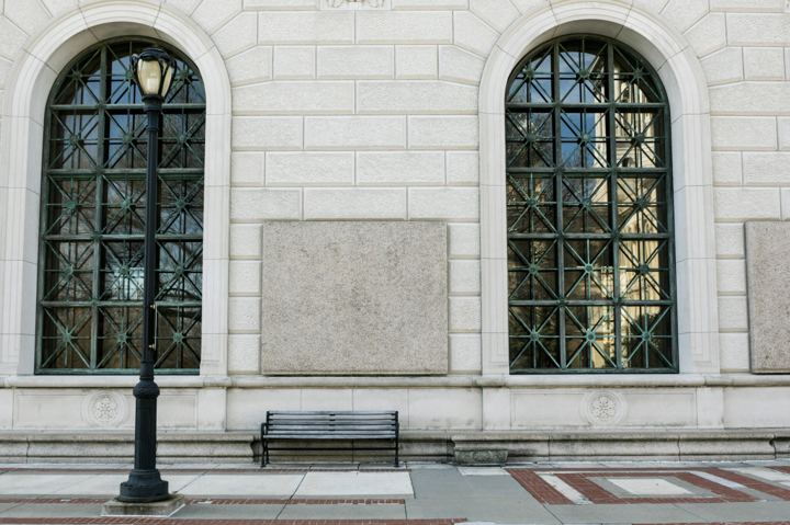 Two tall arched windows with green weathered copper grilles set into a pale limestone wall, an iron lamppost and an empty black bench on the brick sidewalk below.
