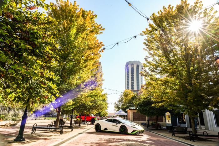 Late day sun flaring through the trees of a downtown plaza, a white supercar parked along the brick walk and a tower rising behind the canopy.