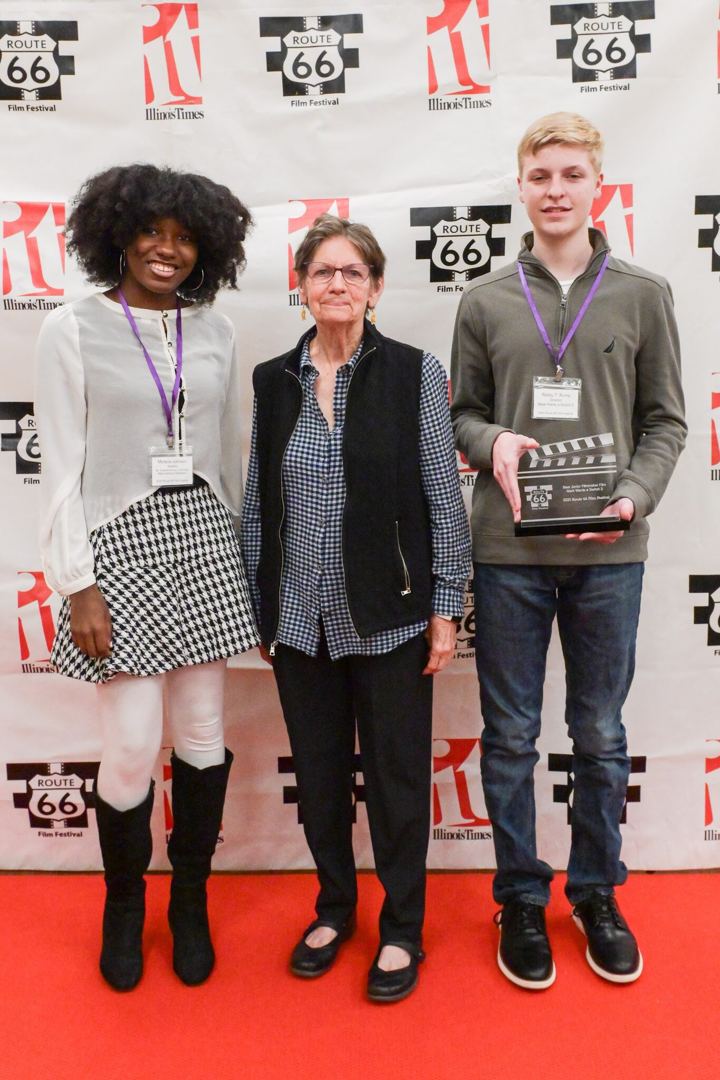 Three people on the red carpet in front of the Route 66 Film Festival backdrop, one of them holding a clapperboard trophy.