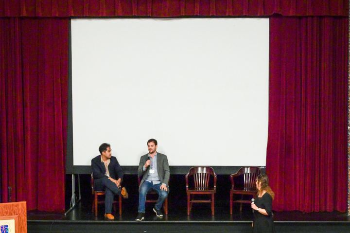 The wide stage at the Hoogland Center for the Arts with the screen up and the red curtain drawn back, two filmmakers seated for a question and answer session and a host standing at the right of the frame.