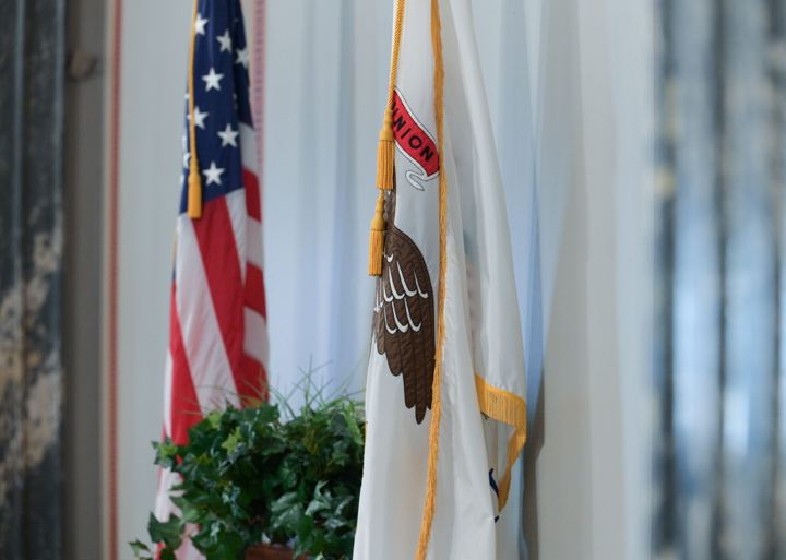 The Illinois state flag hanging on a gold corded pole, the eagle and the UNION ribbon visible in its folds, with the United States flag and a trailing ivy behind it.