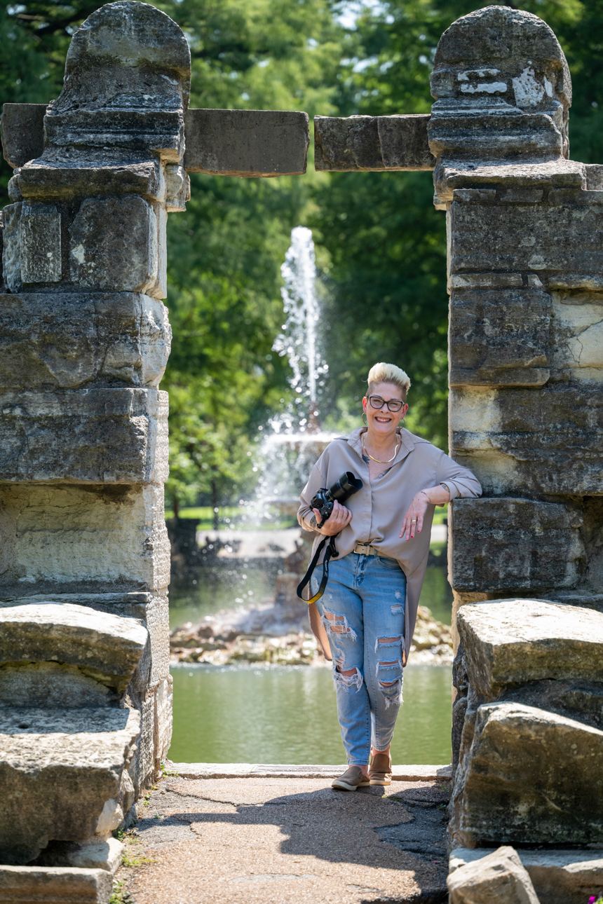 Mindy Pastrovich leaning against one of two weathered stone pillars on a park path, camera in one hand, a tall fountain jetting into a pool behind her and green trees beyond.