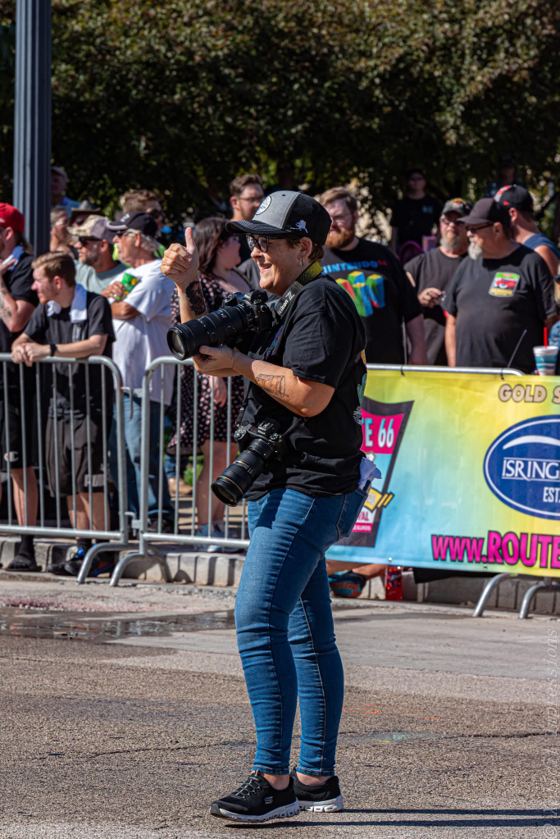 Mindy Pastrovich laughing behind her camera in the middle of a closed street, a crowd lined along the barricades behind her and a Route 66 festival banner at the curb.