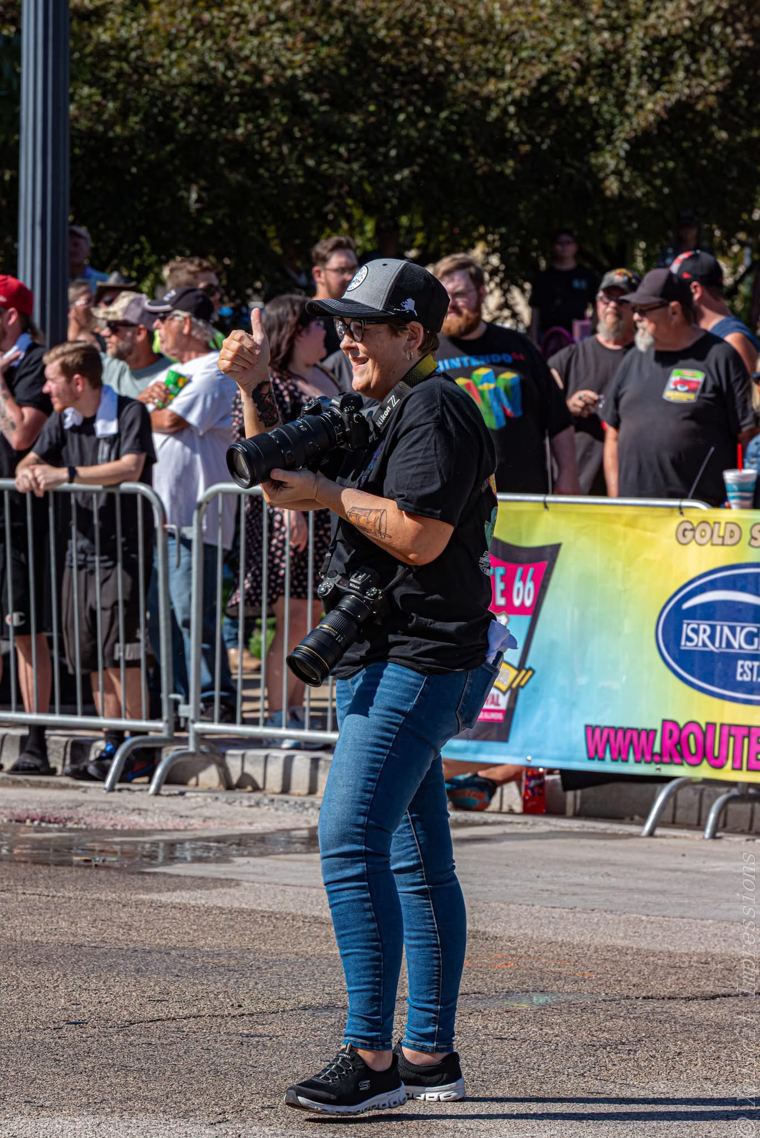 Mindy Pastrovich laughing behind her camera in the middle of a closed street, a crowd lined along the barricades behind her and a Route 66 festival banner at the curb.