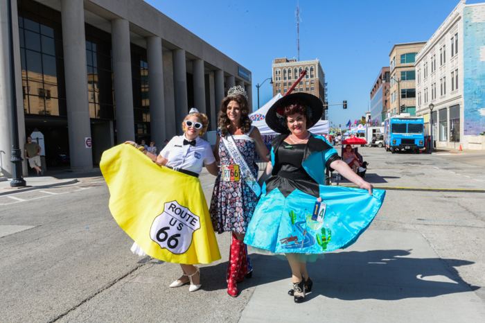 Three women in wide circle skirts, one printed with the Route 66 shield and one wearing a pageant sash, holding their skirts out on a closed downtown street.