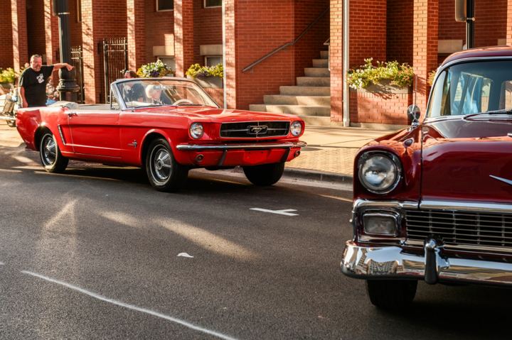 A red Ford Mustang convertible rolling down a downtown street past a brick building, with the chrome grille of a maroon 1956 Chevrolet filling the right side of the frame.