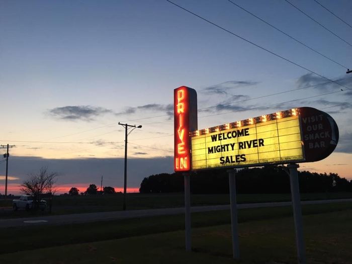 The lit Skyview Drive-In marquee at dusk, red neon spelling DRIVE IN down the pillar, bulbs around a yellow reader board and a panel reading VISIT OUR SNACK BAR, with the last of the sunset along the horizon behind.