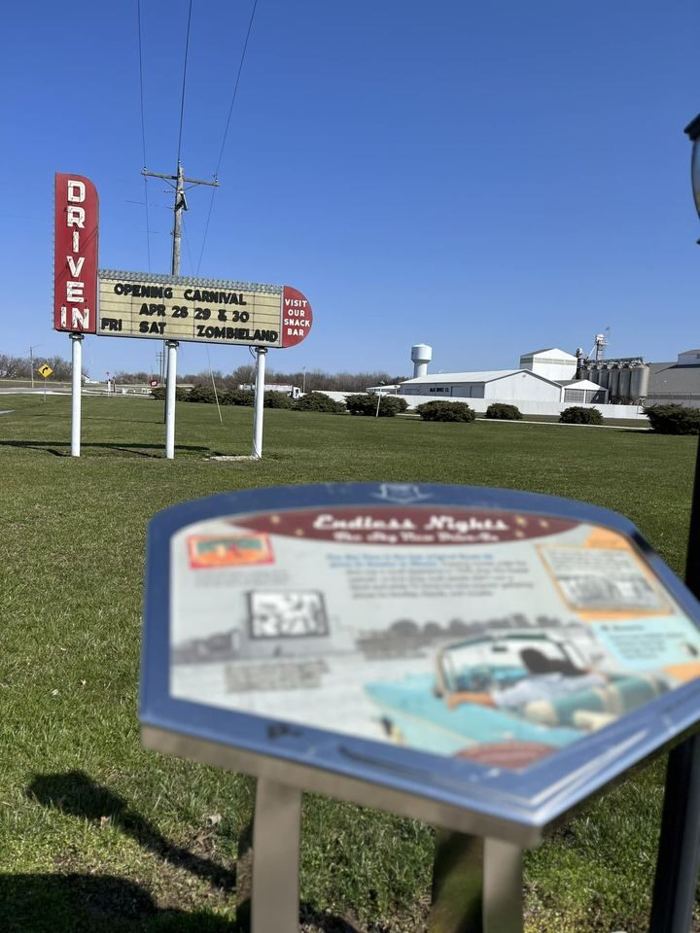 The Skyview Drive-In marquee in daylight reading OPENING CARNIVAL APR 28 29 and 30, with a Route 66 interpretive marker for the drive-in in the foreground and grain bins along the horizon.