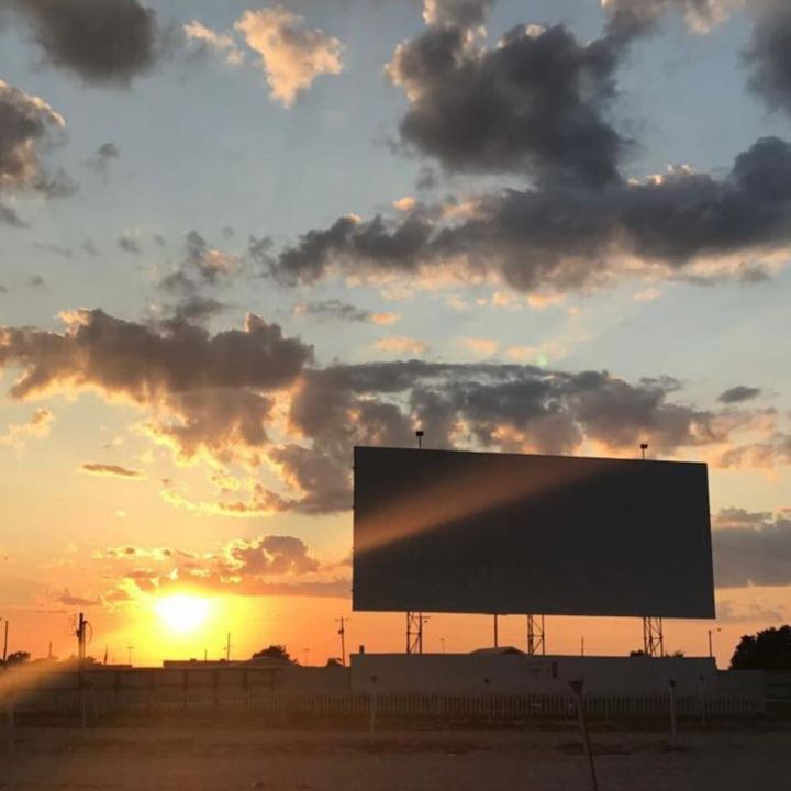 The Skyview Drive-In screen seen from the middle of the gravel lot at sunset, the sun going down at the left edge and broken clouds across the whole sky.