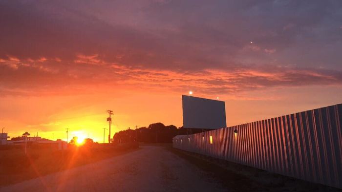 The screen tower of the Skyview Drive-In standing against a red and orange sunset, with the corrugated perimeter fence running back toward the setting sun and the gravel entrance drive alongside.