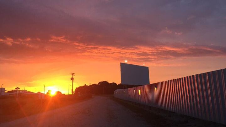 The screen tower of the Skyview Drive-In standing against a red and orange sunset, with the corrugated perimeter fence running back toward the setting sun and the gravel entrance drive alongside.