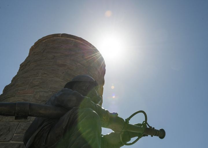 A bronze soldier on a stone monument shot from below into the sun, rifle over one shoulder and a bugle in hand, the light flaring across the figure.